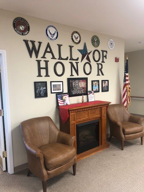 A Wall of Honor display featuring military emblems, photographs, and plaques with an American flag, flanked by two leather chairs and a decorative fireplace.