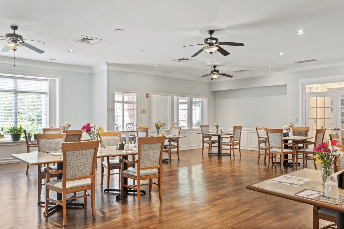 A bright dining room with wooden floors, multiple tables and chairs, ceiling fans, and large windows.
