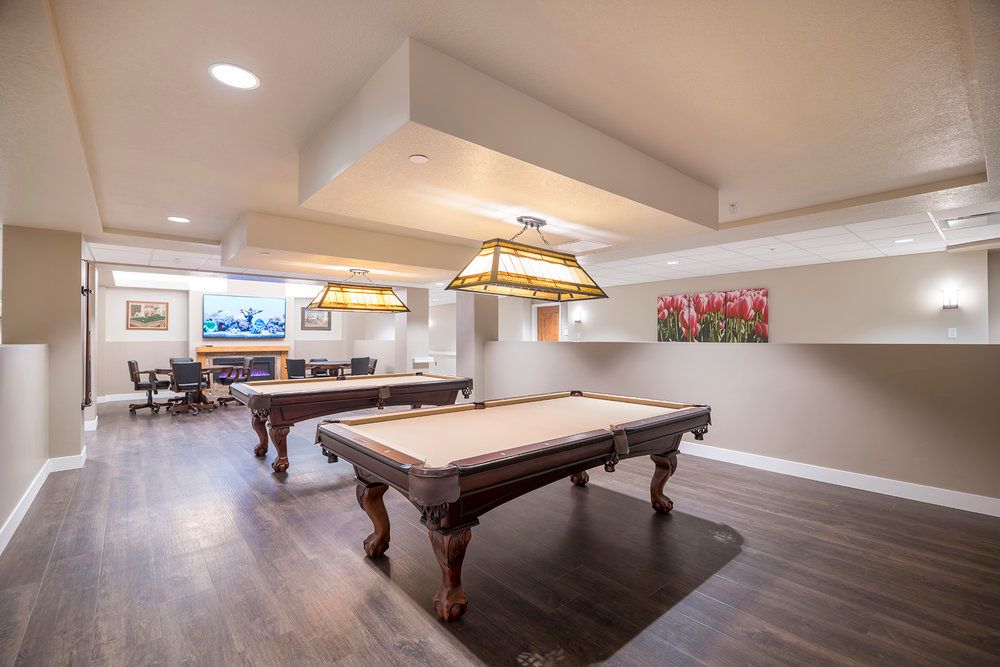 A spacious game room featuring two billiard tables under stylish hanging lights, with wood flooring and a cozy atmosphere.