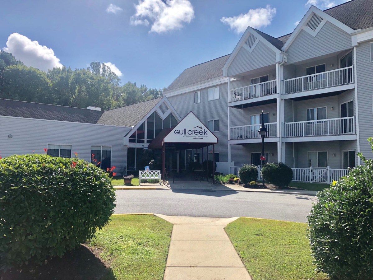 The image shows a residential apartment building with a landscaped entrance on a sunny day.