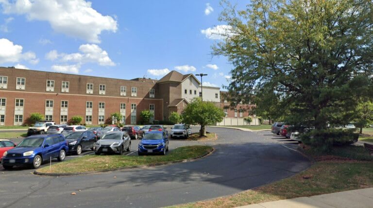A brick building with a parking lot in front, surrounded by trees under a partly cloudy sky.