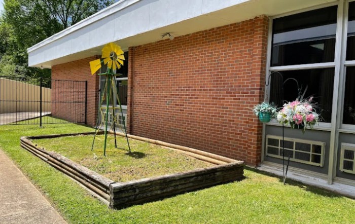 A metal sculpture resembling a sunflower stands in a raised garden bed beside a brick building.