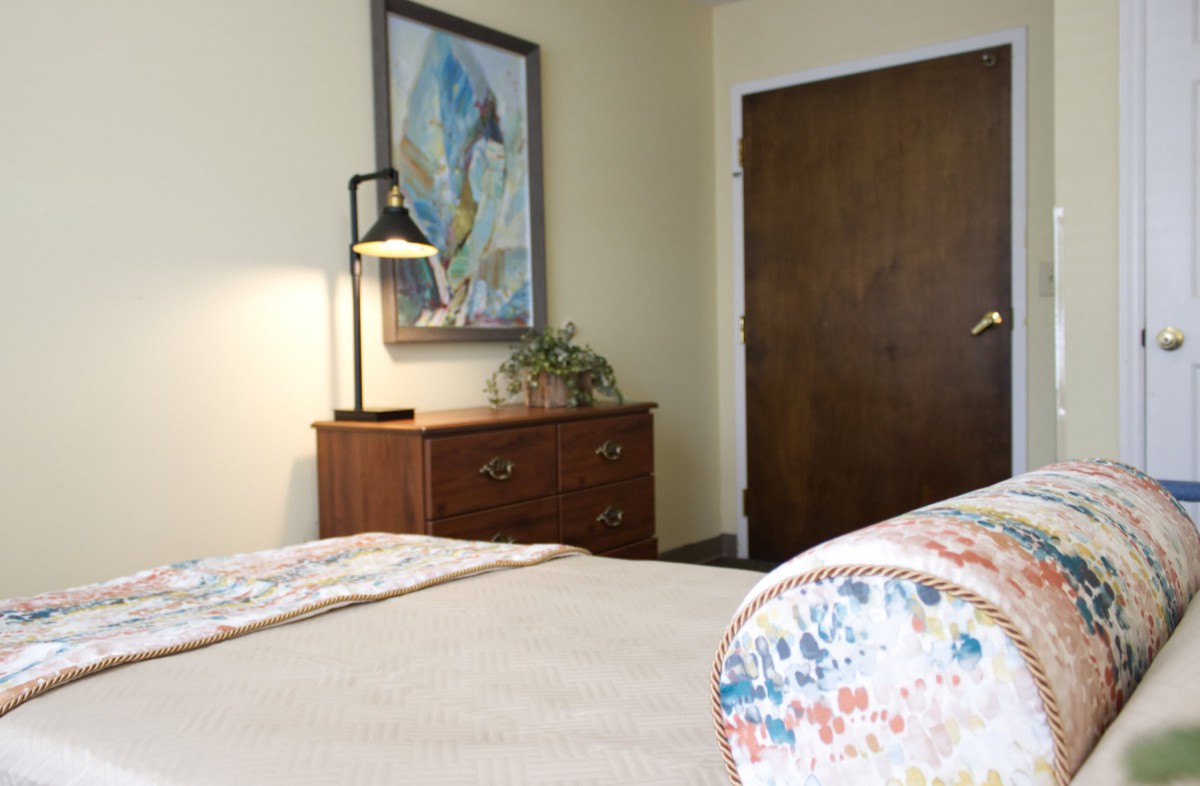 A cozy bedroom with a wooden dresser, an artwork on the wall, and a lamp.