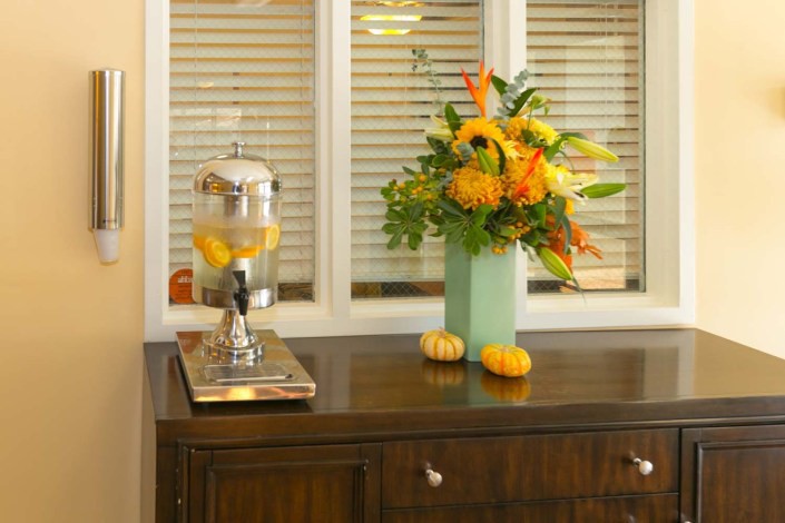 A beautifully arranged autumn-themed interior setup with a floral bouquet, small pumpkins, and a lemon-infused water dispenser on a wooden cabinet.