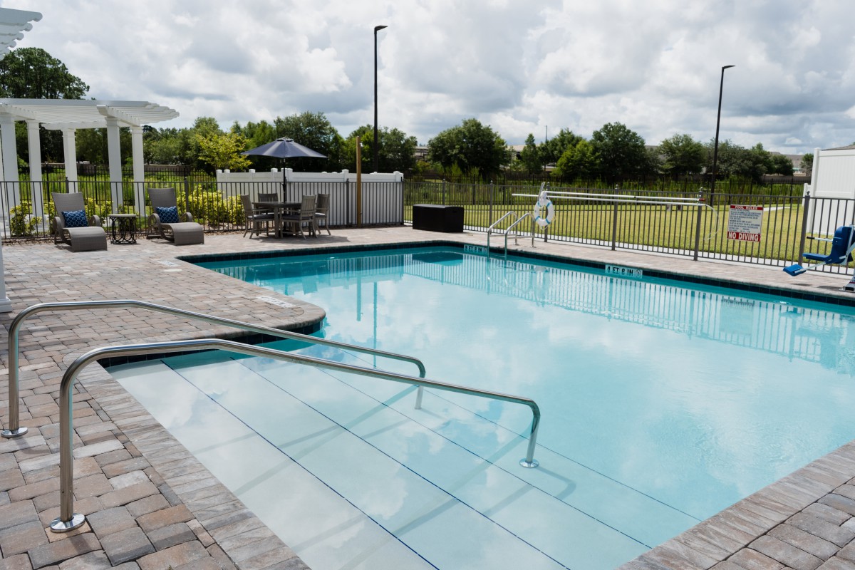 An outdoor swimming pool with patio furniture and a fence under an overcast sky.