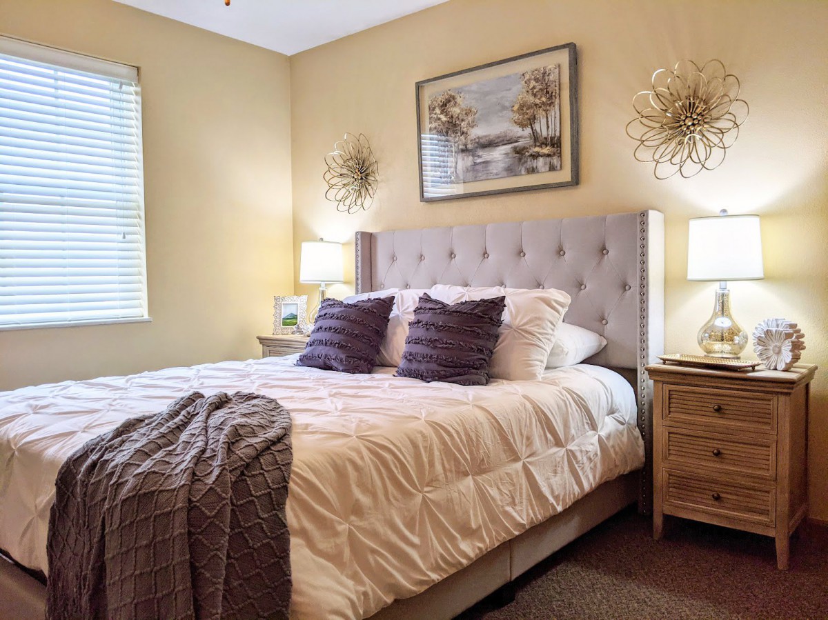 A cozy bedroom with a neatly made bed, featuring tufted headboard and decorative pillows, flanked by nightstands with lamps and wall artwork.