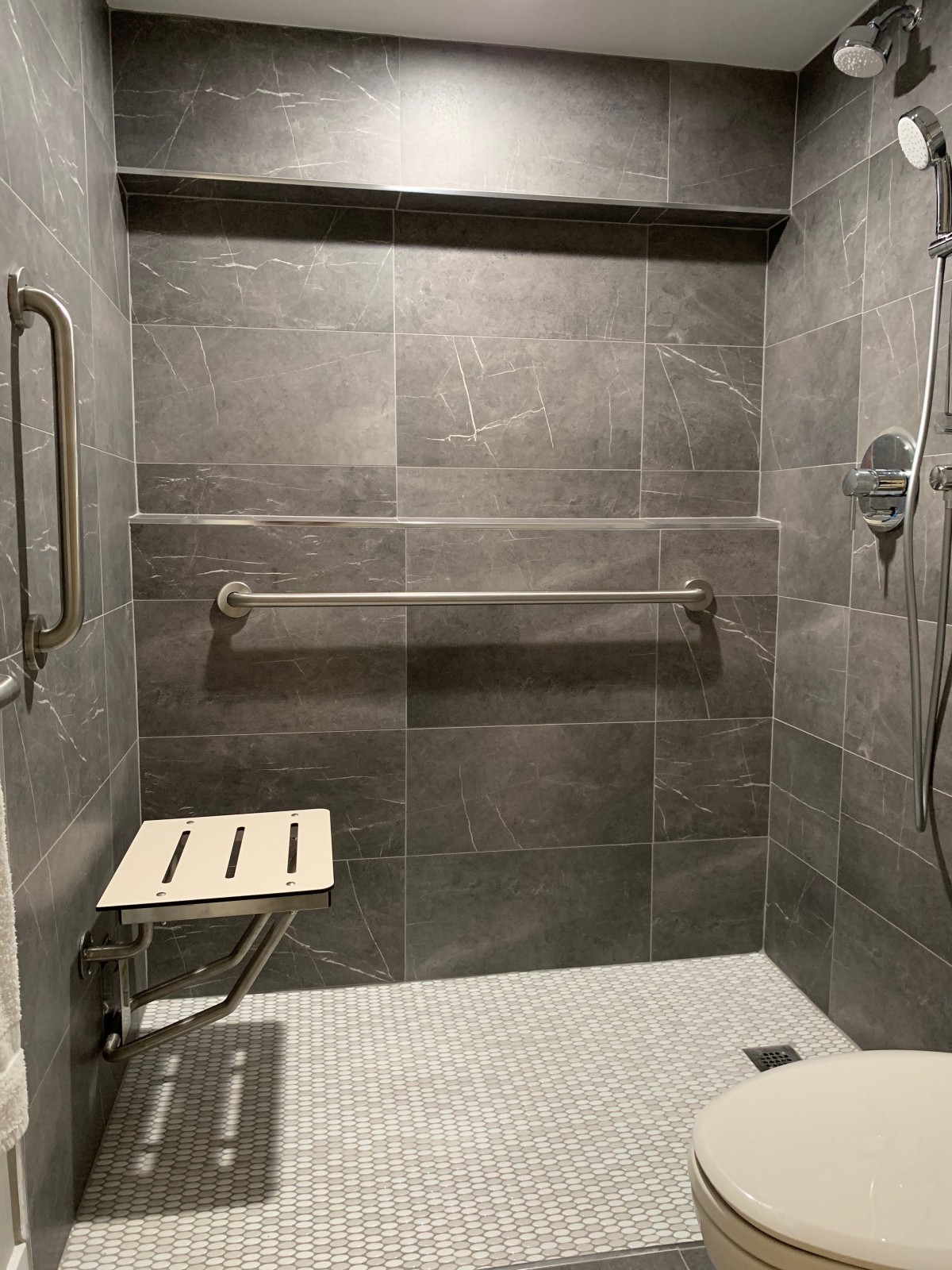 A modern, accessible shower with grey tiles, equipped with a foldable seat and grab bars for safety.