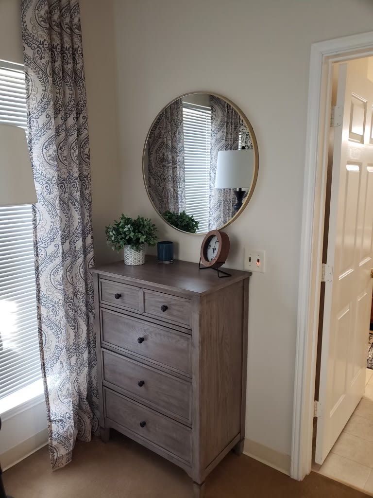 A bedroom corner featuring a wooden dresser with a round mirror, a small plant, and decorative items. Natural light streams in through the window with patterned curtains.