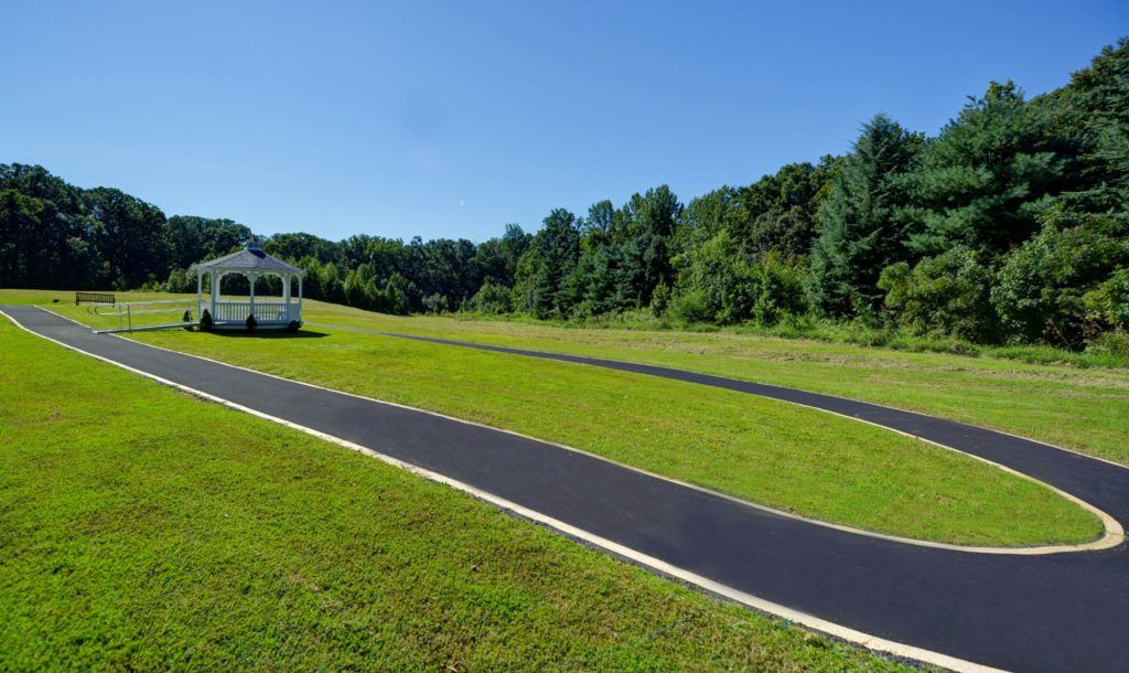 A paved walkway curves around a well-maintained lawn with a gazebo in the background, surrounded by trees under a clear blue sky.