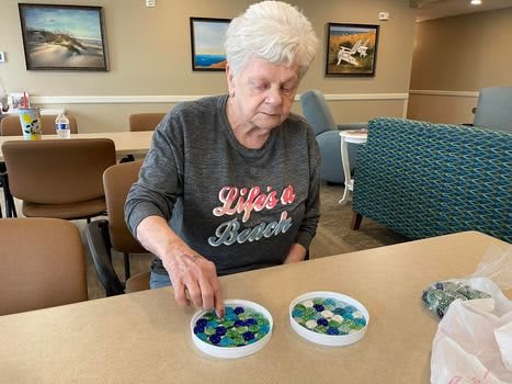 An elderly woman engages in a crafting activity, arranging beads on a table.