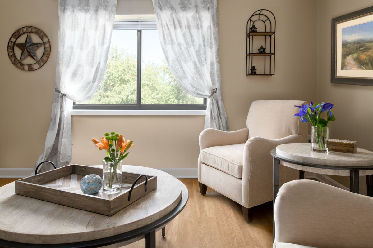 A cozy living room with a comfortable armchair and a coffee table adorned with flowers, set against a sunny window with sheer curtains.