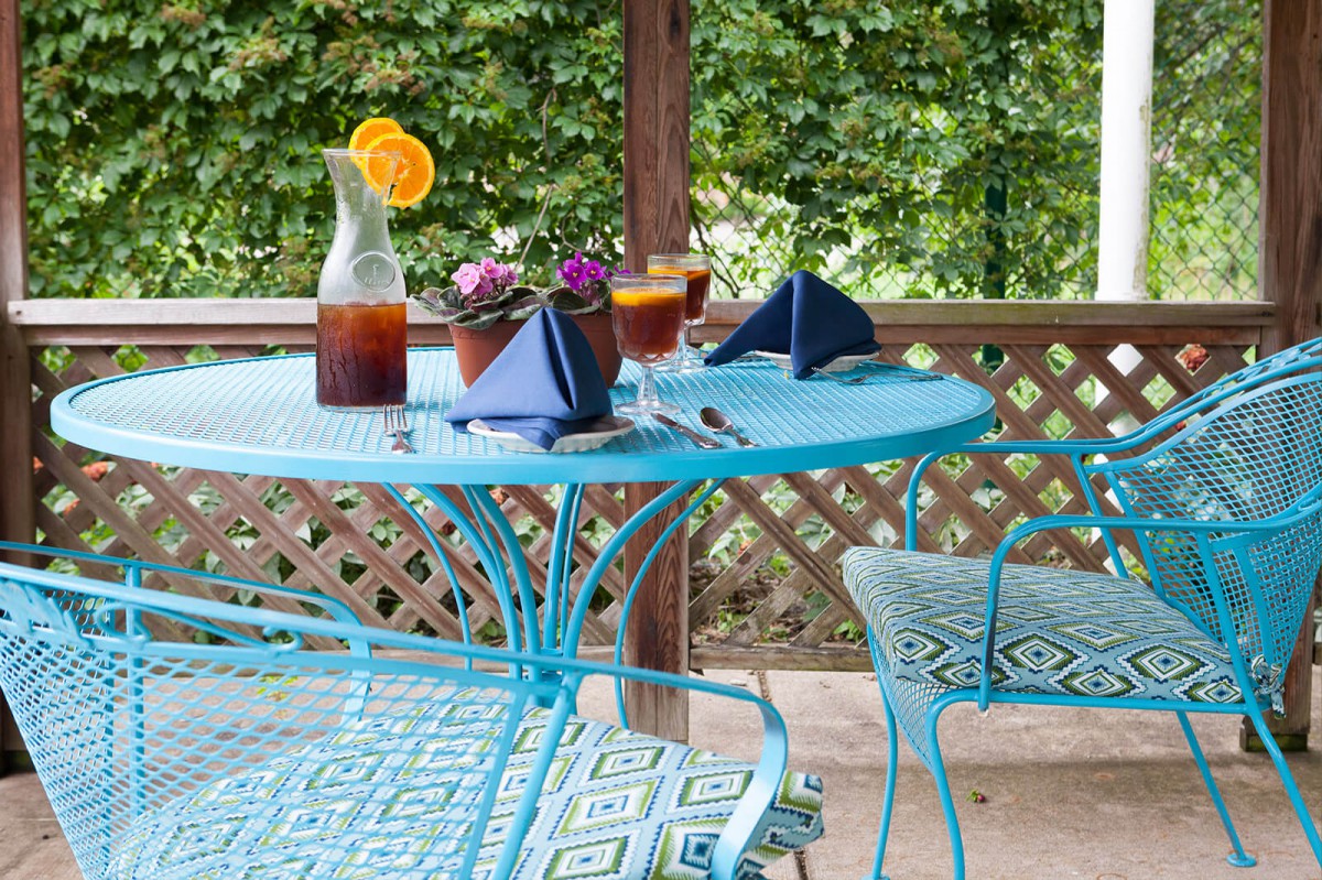 A cozy outdoor table setting with blue furniture, featuring iced tea and decorative plants against a garden backdrop.