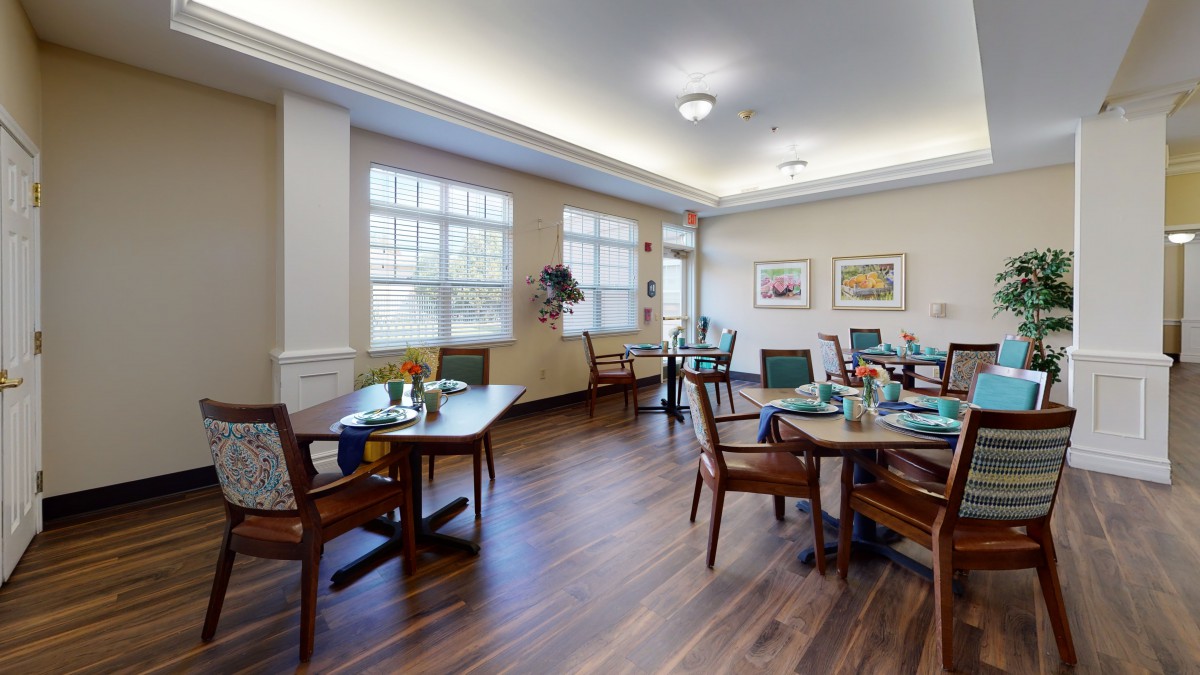 A cozy dining room with neatly arranged tables and chairs, featuring large windows and warm lighting.