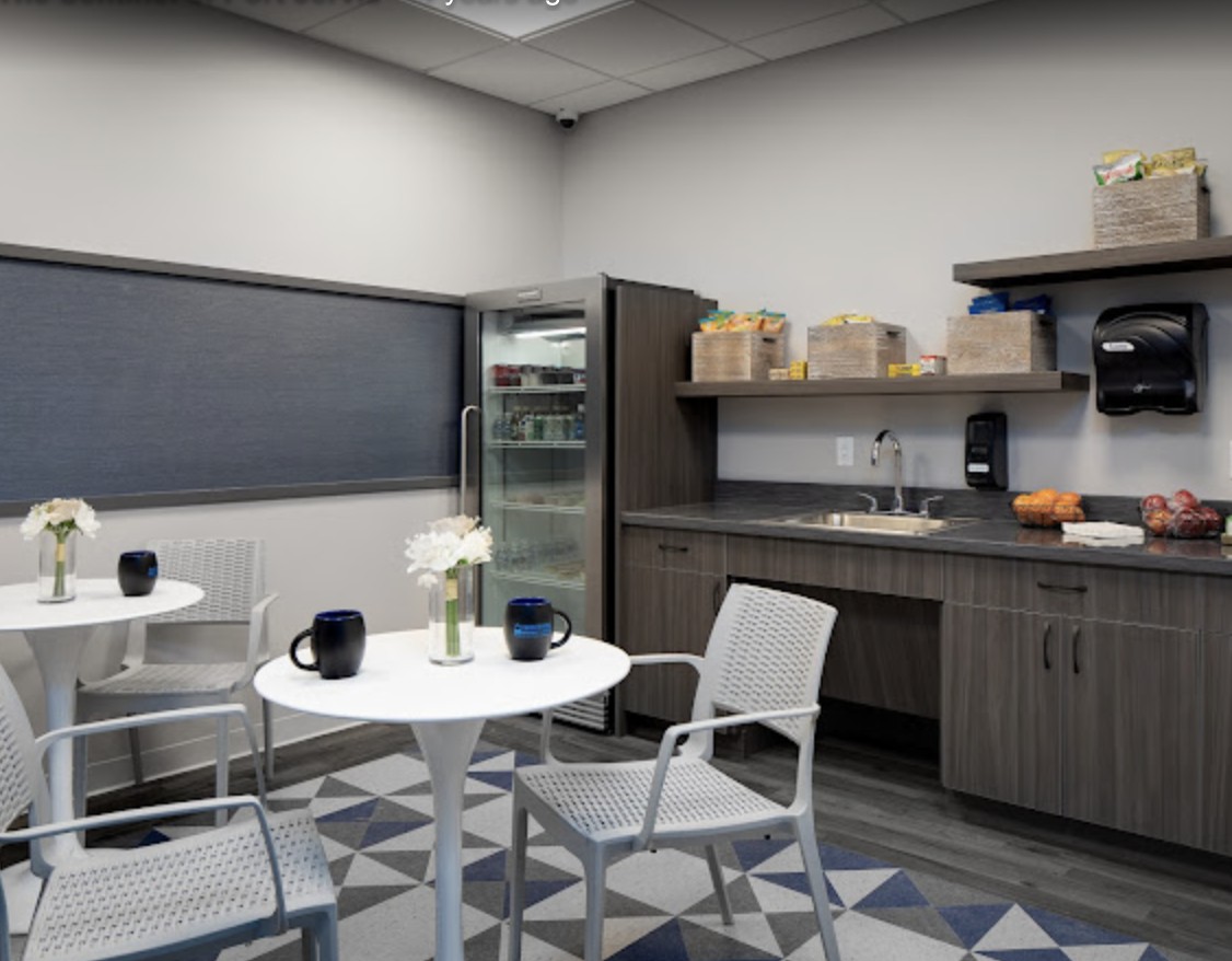 A modern break room with a small kitchenette, featuring tables, chairs, and a refrigerator.