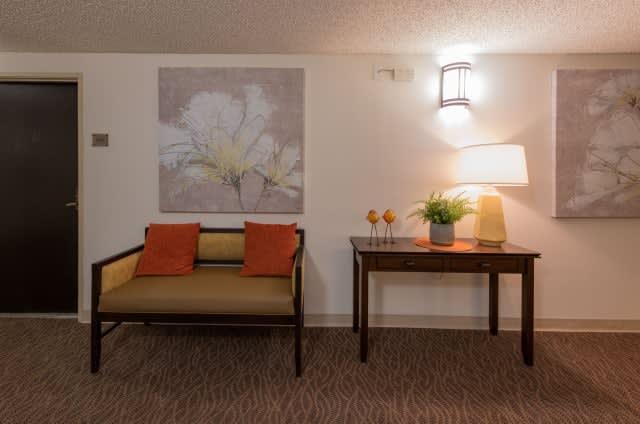 A cozy sitting area featuring a small bench with orange cushions, a decorative table with a lamp and plants, and floral artwork on the wall.