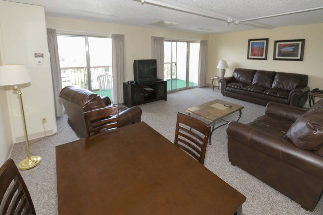 A spacious living room with brown leather sofas, a dining table, and a television, featuring large windows that let in natural light.