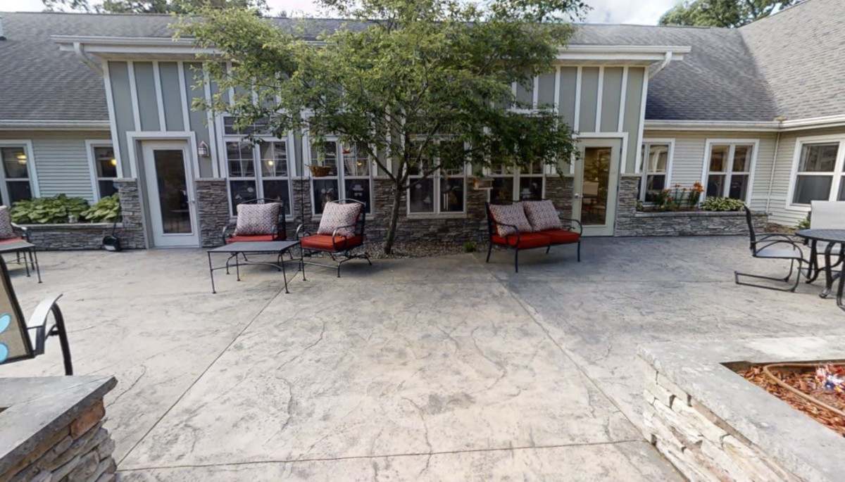 A cozy patio area with cushioned chairs under a tree, in front of a building with multiple windows.