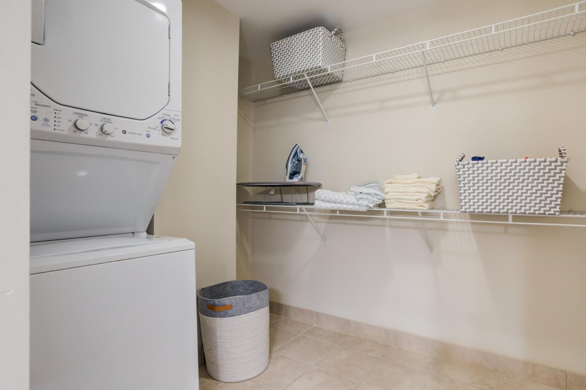 Neat and organized laundry room with a stacked washer and dryer, ironing board, and clean shelves.
