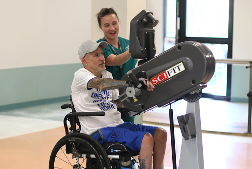 A patient in a wheelchair engages with exercise equipment during a physical therapy session, assisted by a healthcare professional.