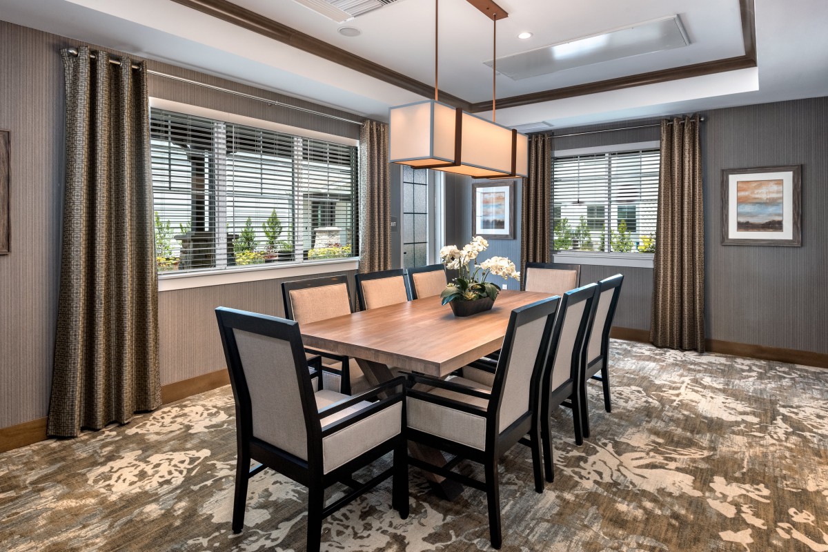 A modern dining room featuring a large wooden table surrounded by upholstered chairs, with ample natural light from the windows and an elegant overhead lighting fixture.