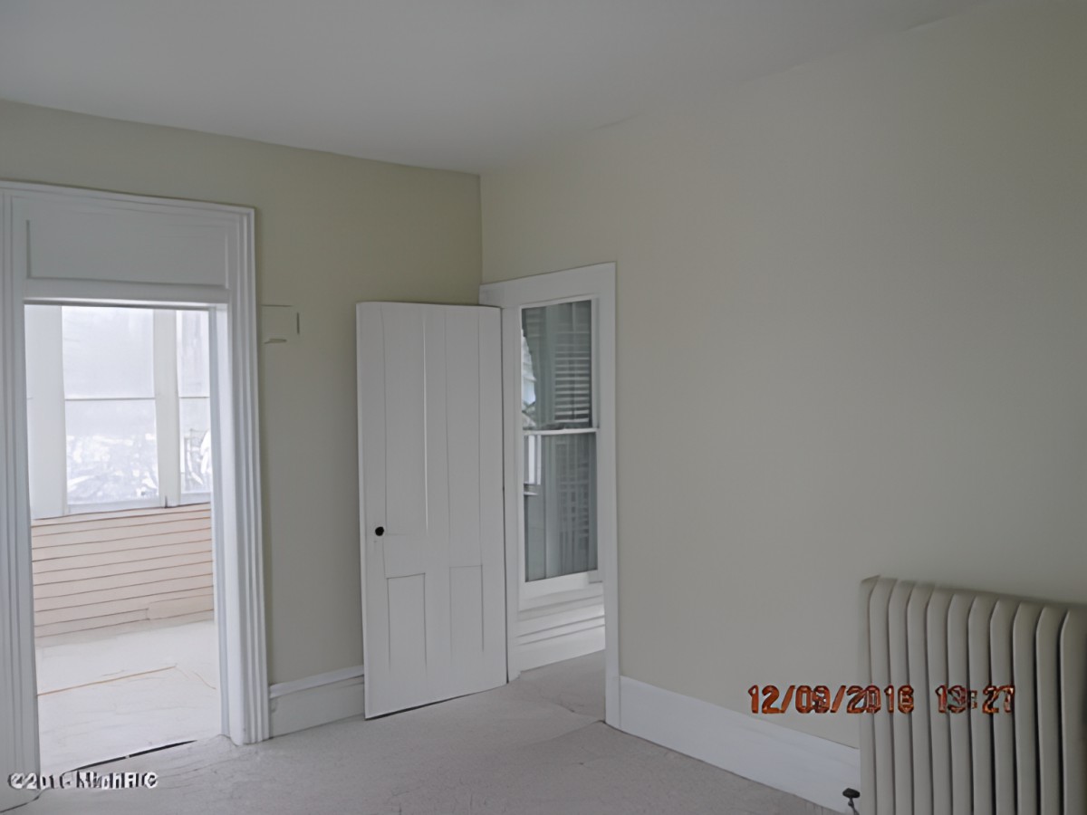 An empty room with light-colored walls, a door leading to another space, and a radiator beneath the window.
