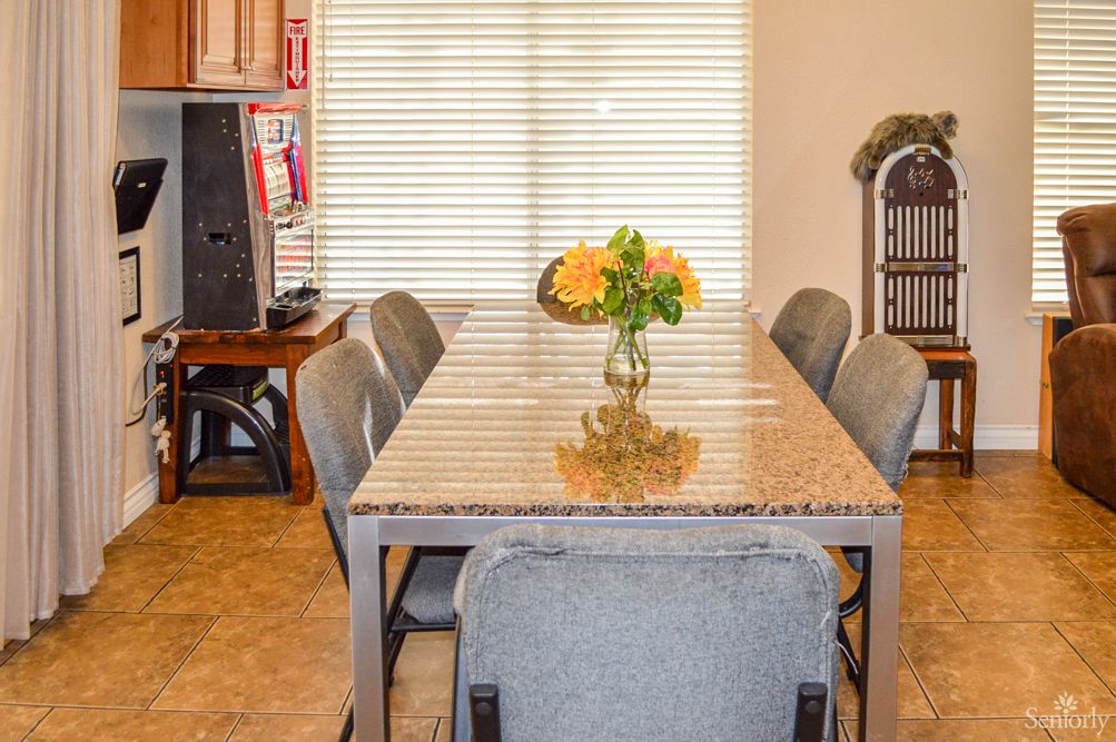 A dining area featuring a long table with gray chairs and a vase of flowers as the centerpiece, complemented by window blinds in the background.