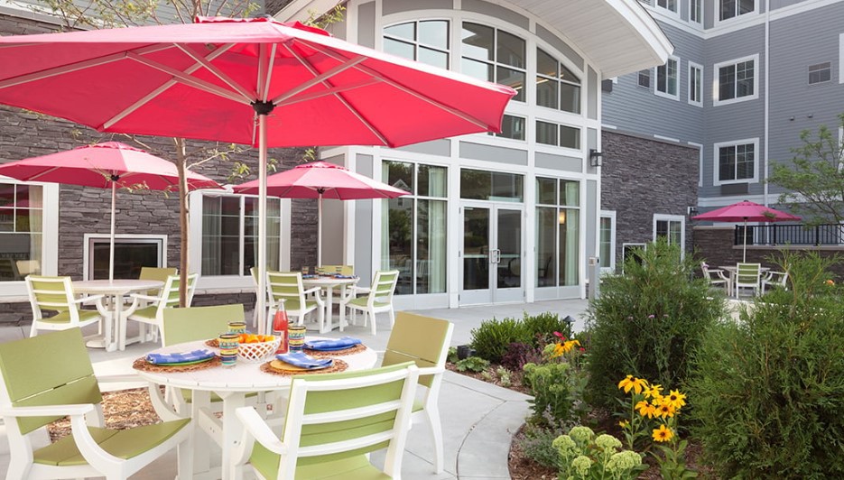 A charming patio area with tables, chairs, and pink umbrellas surrounded by a garden in front of a modern building.
