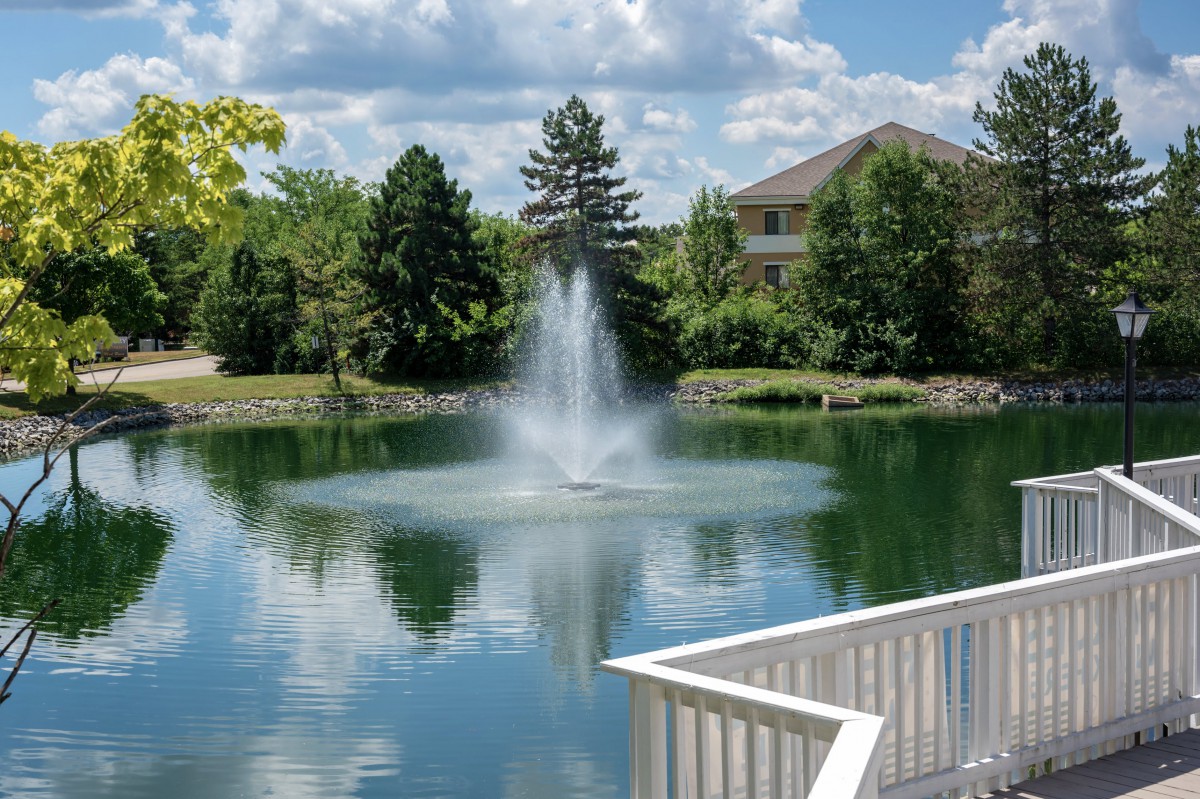 A serene view of a fountain in the center of a small lake, surrounded by trees and a house, with clouds reflecting on the water's surface.