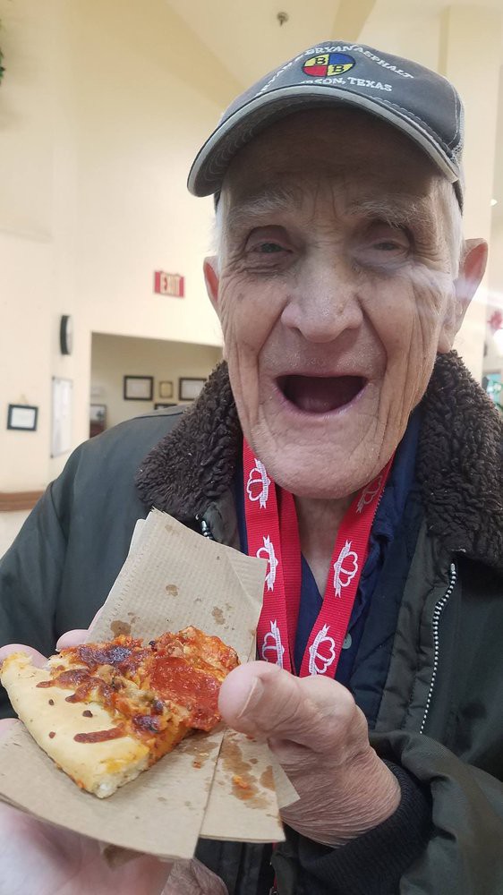 An elderly man joyfully holding a slice of pizza indoors.