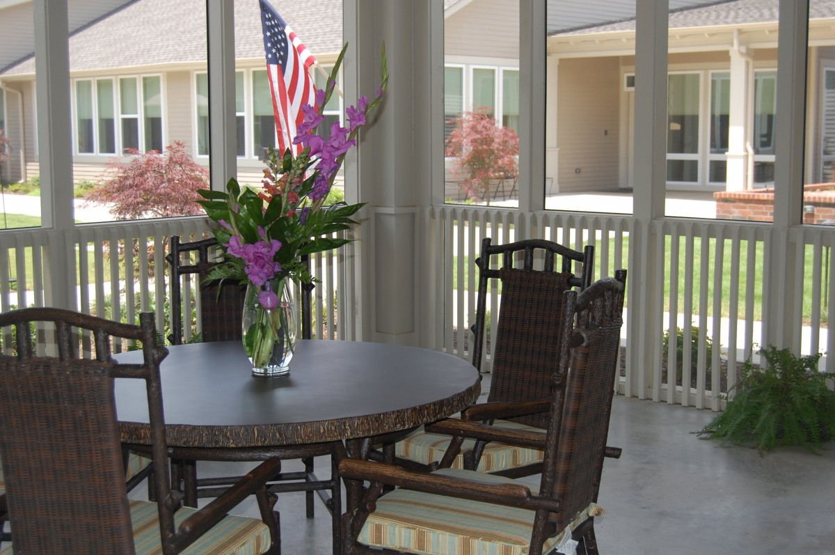 A cozy patio setting with a table, chairs, and a vase of flowers, overlooking an outdoor space with an American flag in the background.