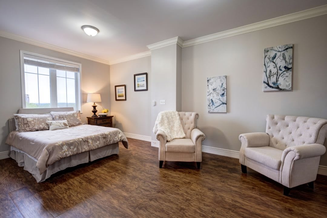 A cozy bedroom with a bed, two armchairs, and decorative artwork on the walls, featuring a wooden floor and natural lighting.