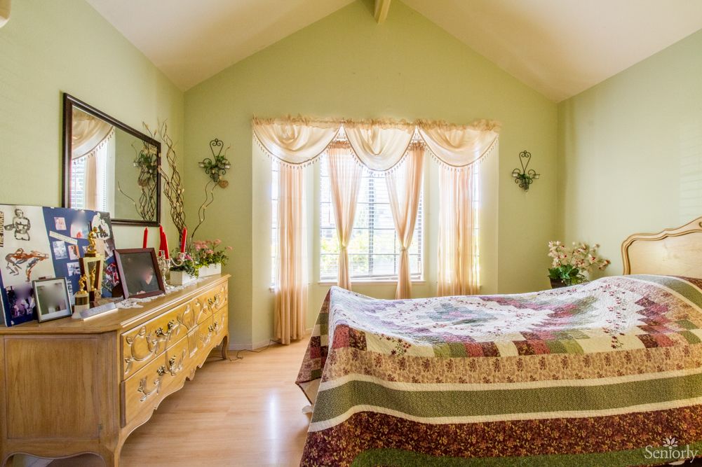 A cozy bedroom with soft green walls, a large window adorned with curtains, and a quilted bedspread on a neatly made bed.