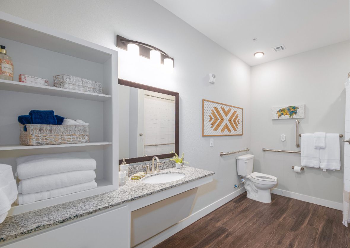 A modern bathroom with a spacious sink area, mounted mirror, toilet, neatly arranged towels on shelves, and stylish wall art.
