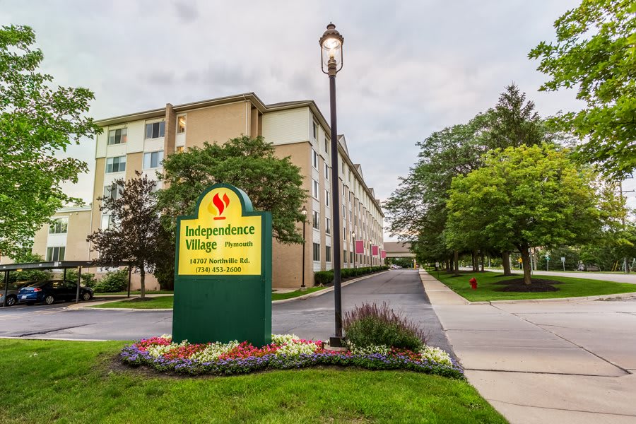 A suburban building with a sign for 'Independence Village' surrounded by trees and flowers along a driveway.