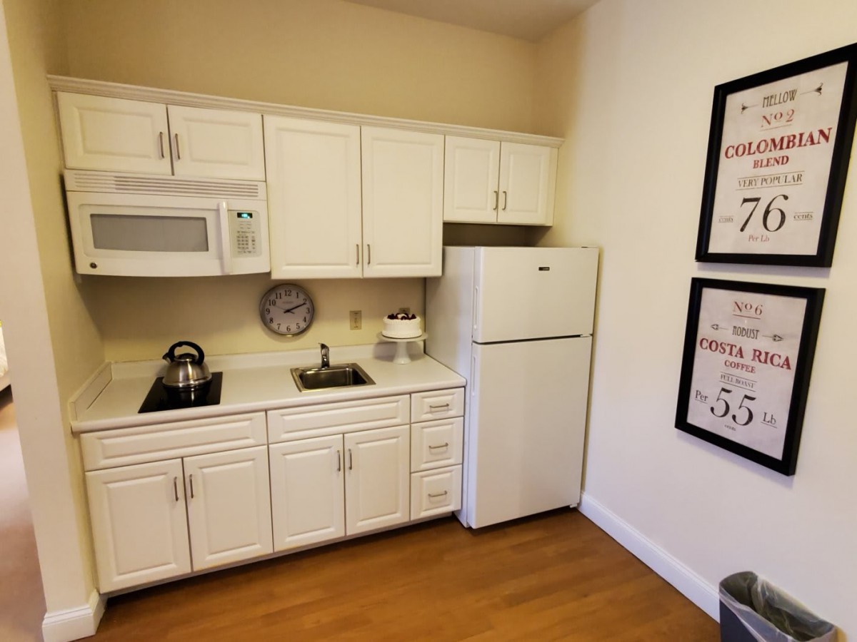 A compact kitchen with white cabinets, equipped with a refrigerator, microwave, and small sink. The space features a kettle on the countertop and framed wall art.