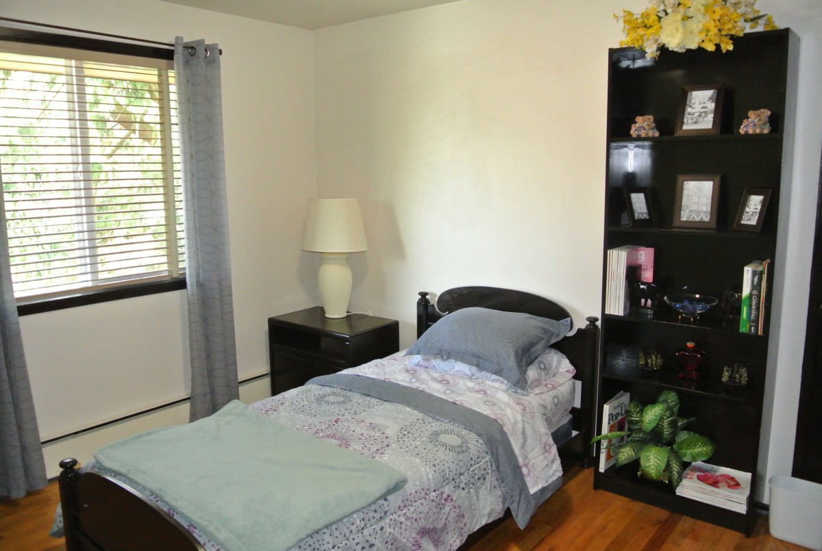 A minimalist bedroom with a single bed, a lamp on a nightstand, and a bookshelf.