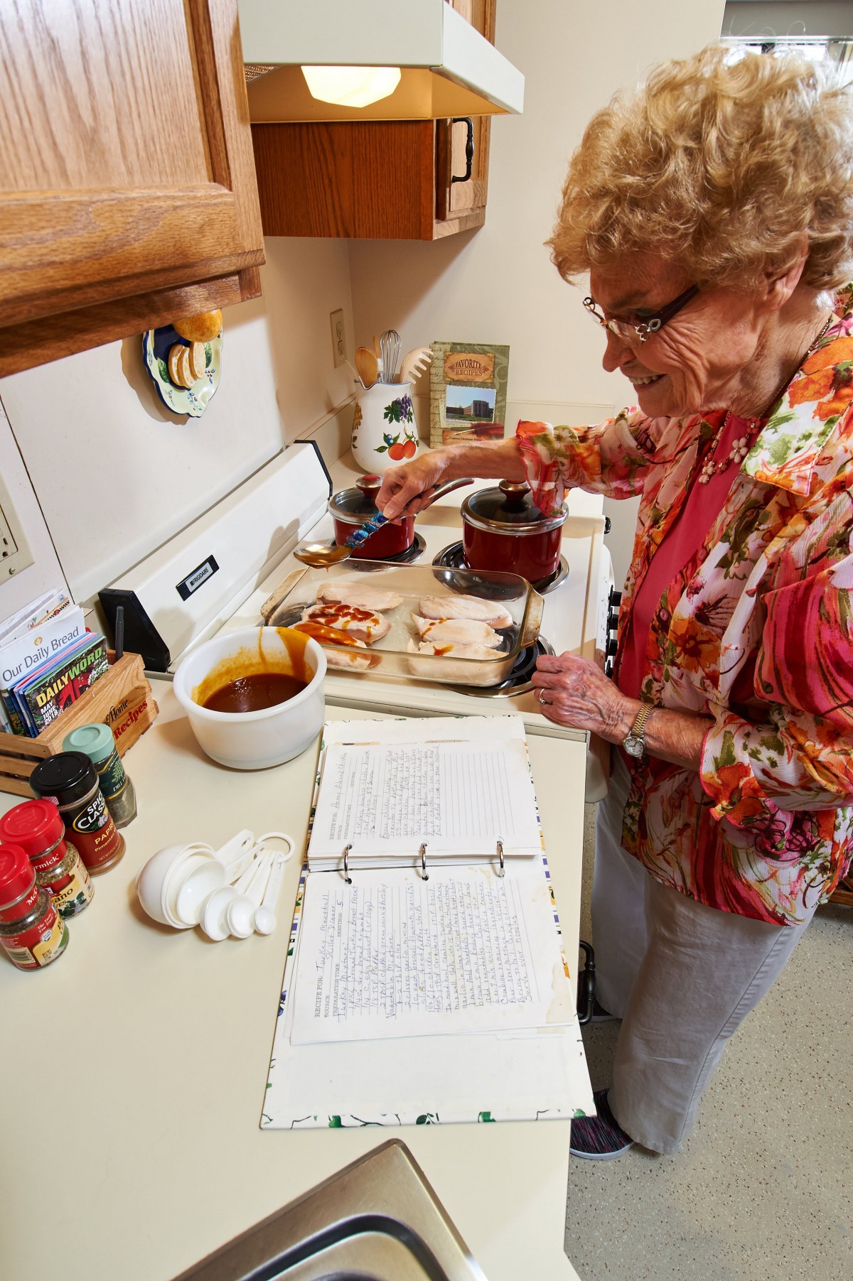 An elderly woman preparing a meal in the kitchen while referencing a recipe book.