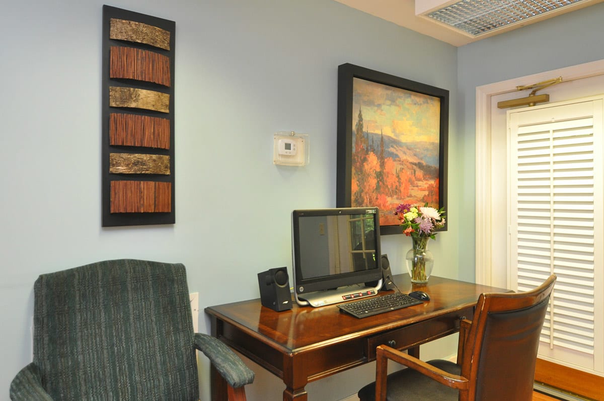 A small office space with a computer on a wooden desk, surrounded by artwork and flowers.