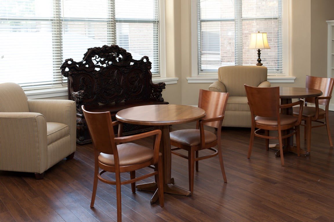 A cozy living room area with wooden chairs and a round table, surrounded by soft armchairs and lit by natural light through large windows.