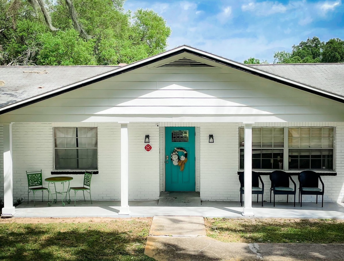 A charming single-story house with a white facade and a teal front door, featuring outdoor seating on the porch.
