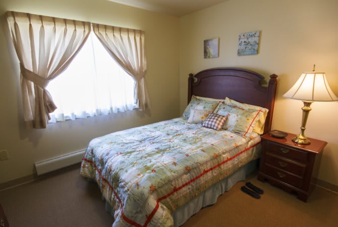 A cozy bedroom with a neatly made bed, floral bedding, and curtains allowing natural light to fill the room.