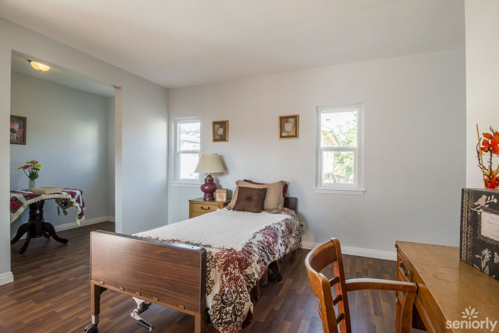 A cozy bedroom with a wooden bed, chairs, and a lamp on a side table, featuring decor pieces and natural light from two windows.
