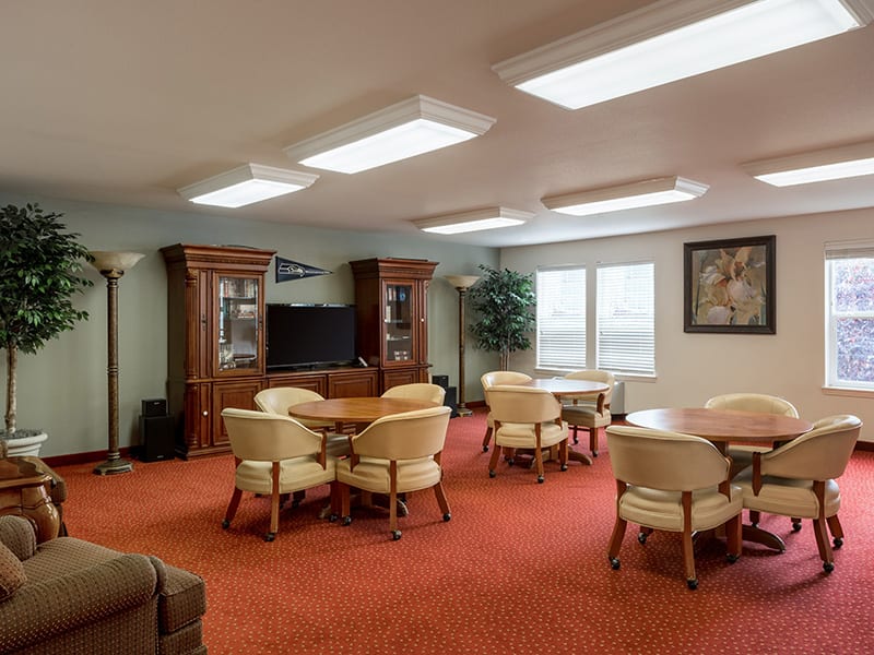 A spacious meeting room with tables and chairs on a red carpeted floor, featuring wooden cabinets and a television.
