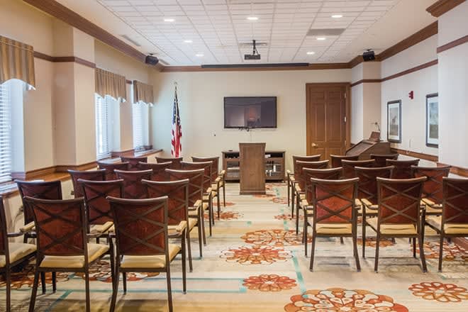 A well-organized conference room with rows of chairs facing a podium and a projection screen, featuring an American flag and large windows.