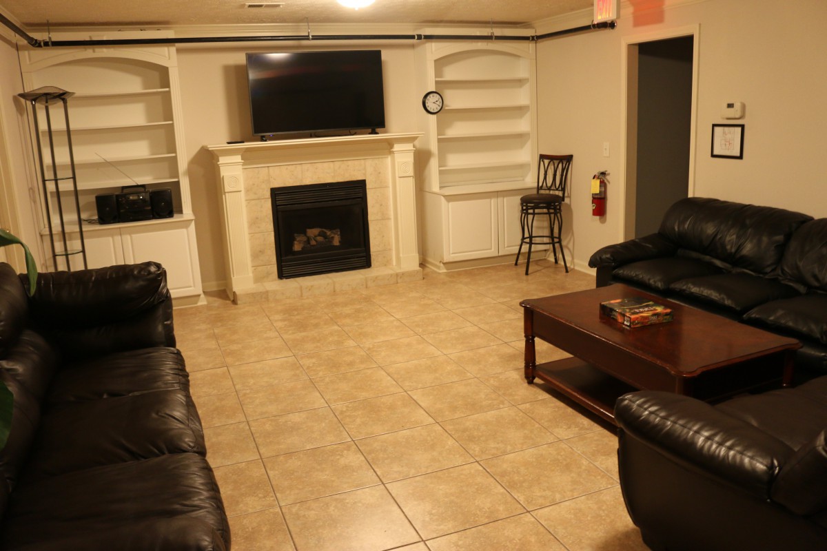 A cozy living room with a television mounted above a fireplace, surrounded by shelves, featuring leather couches and a wooden coffee table on a tiled floor.