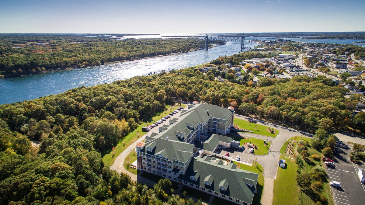 Aerial view of a residential building near a river and bridge surrounded by lush greenery.