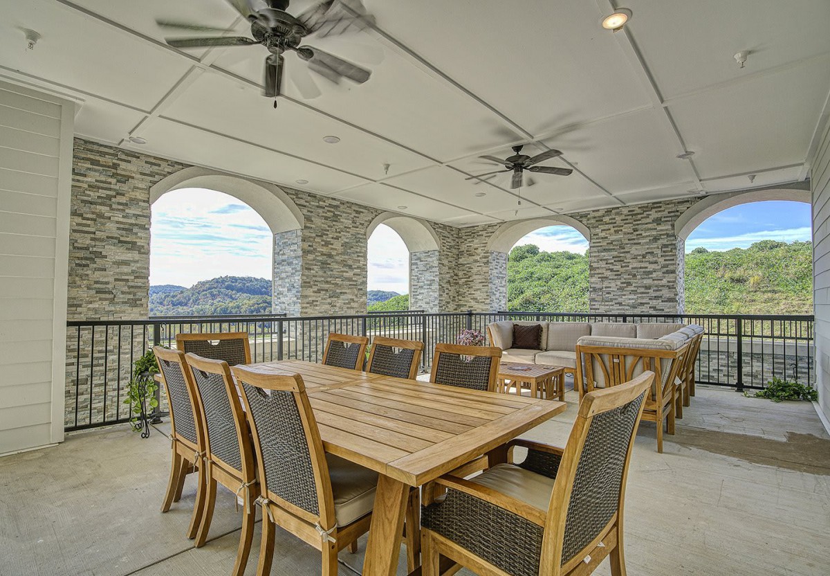 A spacious outdoor patio with wooden dining tables and chairs, featuring stone arches and ceiling fans, offering a scenic view.