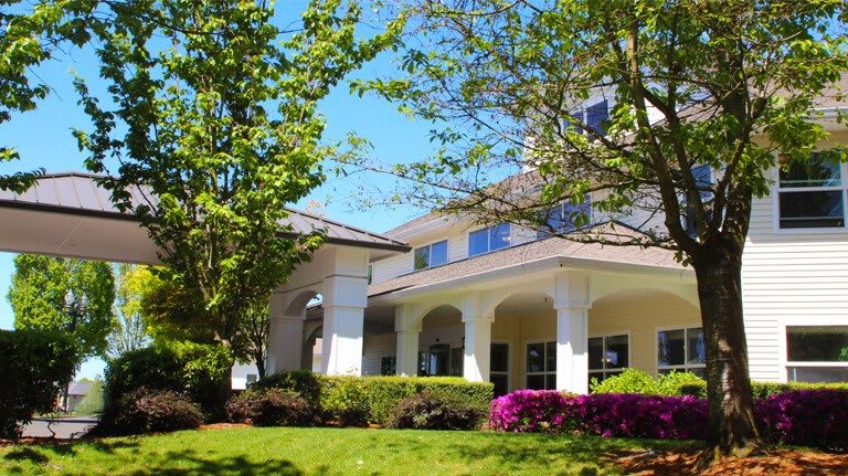 A sunny view of a building surrounded by trees and flowering bushes.