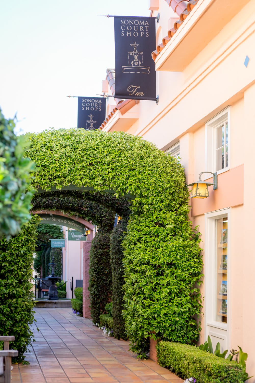 A lush green archway leads to a pathway beside a peach-colored building adorned with signage.