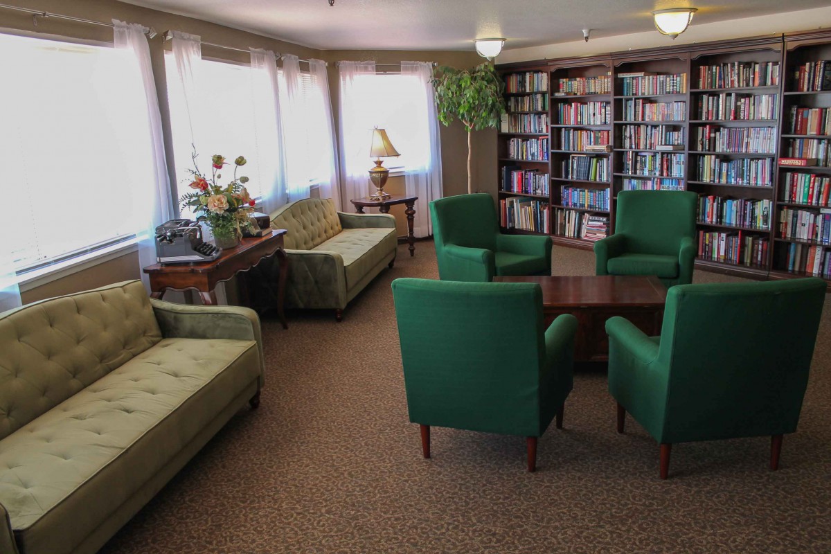 A cozy library room featuring green armchairs and sofas, surrounded by bookshelves and illuminated by natural light through large windows.